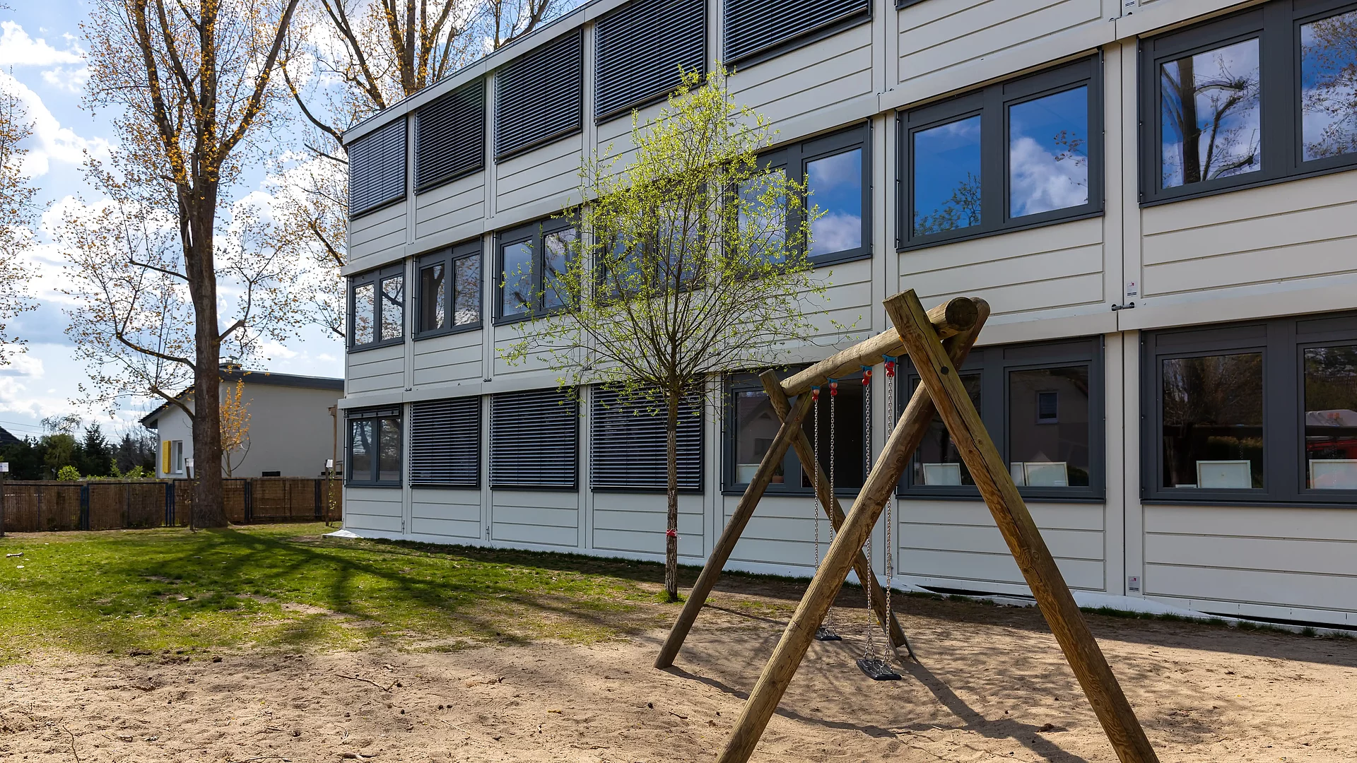 Modulgebäude mit hellem Außenbereich, Sandplatz und Holzschaukel im Vordergrund