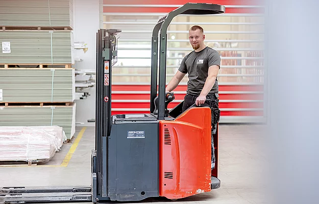 Ein Mann fährt einen orange-schwarzen Gabelstapler in einer Lagerhalle. Im Hintergrund sind gestapelte Paletten und eine rot-weiße Wand zu sehen. Text auf dem Gabelstapler: "RICHTER Fördertechnik + Co.".