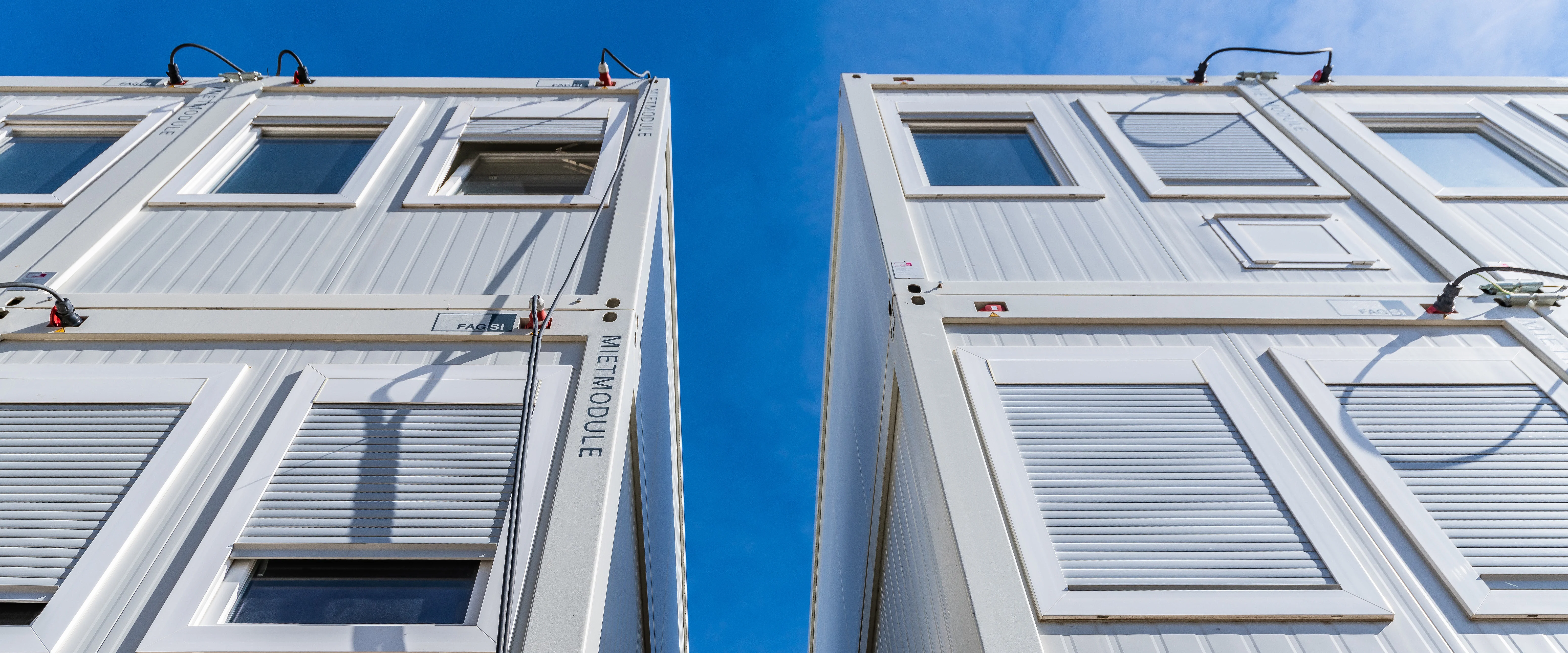 Zwei weiße Mietcontainer stehen dicht nebeneinander und ragen nach oben; Fenster mit teilweise geschlossenen Rollläden, Kabel und Beschriftung "MIETMODULE" sichtbar; klarer blauer Himmel im Hintergrund.