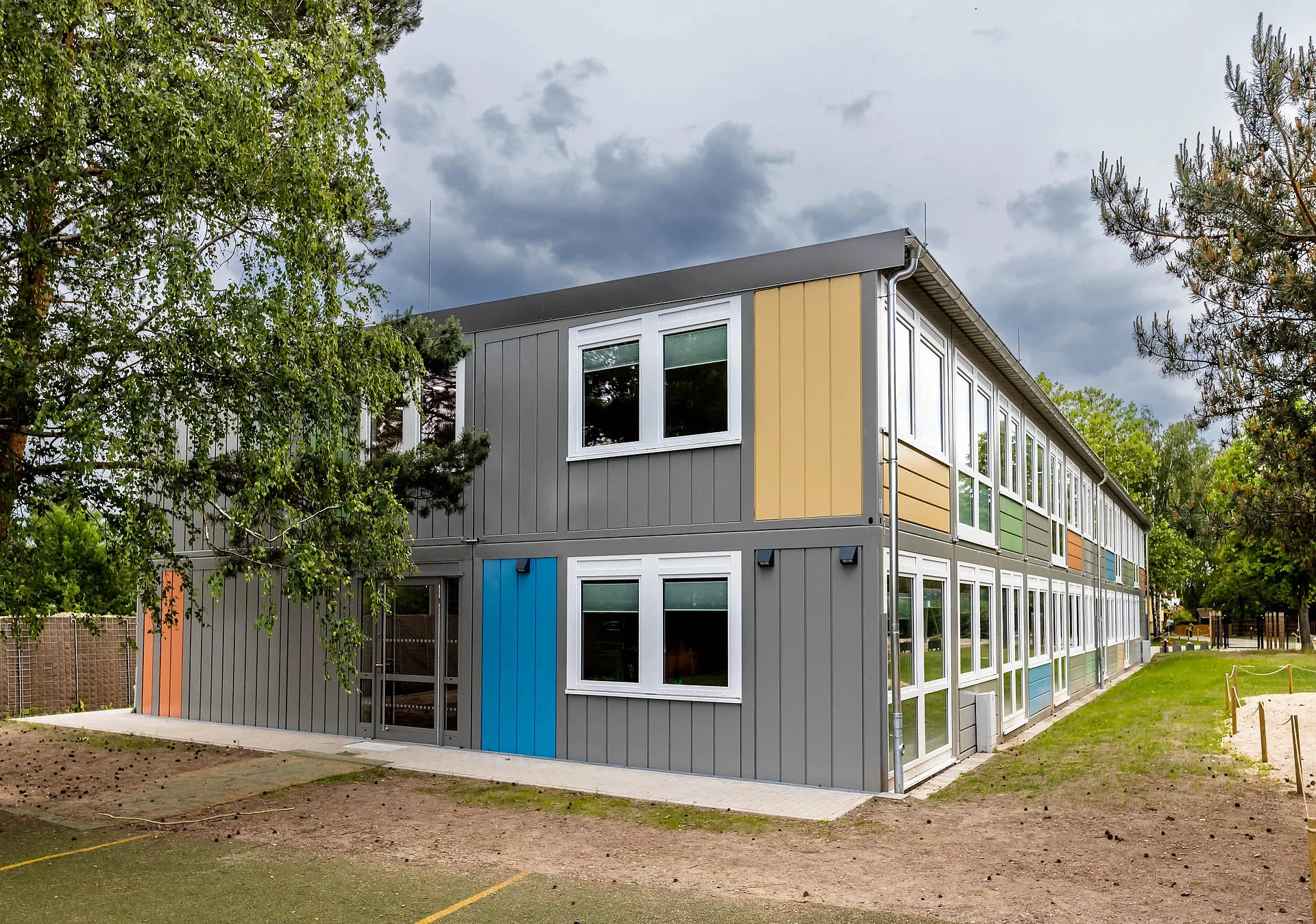 Zweigeschossiges modernes Modulgebäude mit grauer Fassadenverkleidung und farbigen Akzenten in Blau, Gelb, Grün und Orange. Das langgestreckte Gebäude verfügt über zahlreiche große Fenster und ist von Bäumen sowie einer Grünfläche umgeben. Im Vordergrund befindet sich ein Eingangsbereich mit verglaster Tür, während dunkle Wolken den Himmel im Hintergrund prägen.