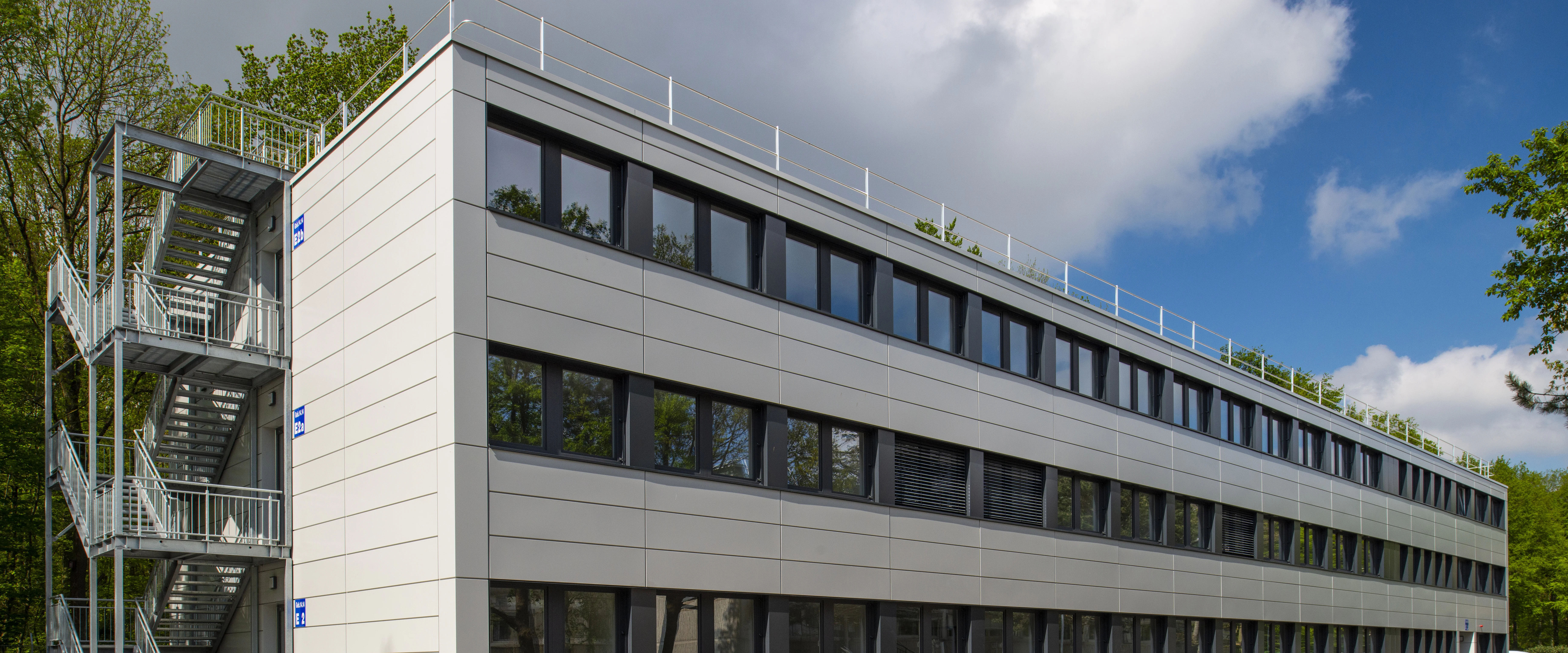 Modernes Bürogebäude mit drei sichtbaren Etagen; lange Reihen dunkler Fenster reflektieren Bäume; hellgraue Fassadenpaneele; Flachdach mit Geländer, blauer Himmel und Wolken im Hintergrund.
