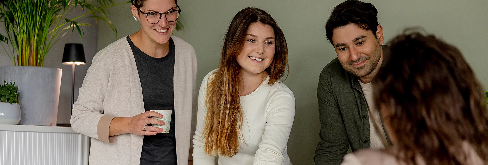 Drei Personen lächeln, während sie diskutieren. Eine Frau hält eine Tasse. Im Hintergrund sind eine Pflanze, eine Lampe und ein Schrank zu sehen.
