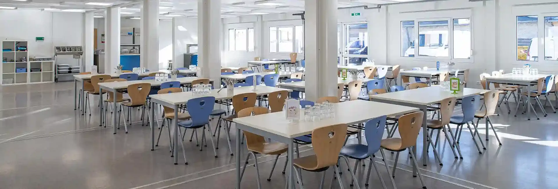 Intérieur lumineux d’une cantine scolaire modulaire avec de nombreuses tables et chaises, aménagée pour accueillir les élèves dans un environnement fonctionnel et confortable. Les grandes fenêtres favorisent l’apport de lumière naturelle et illustrent la qualité des espaces créés en construction modulaire.