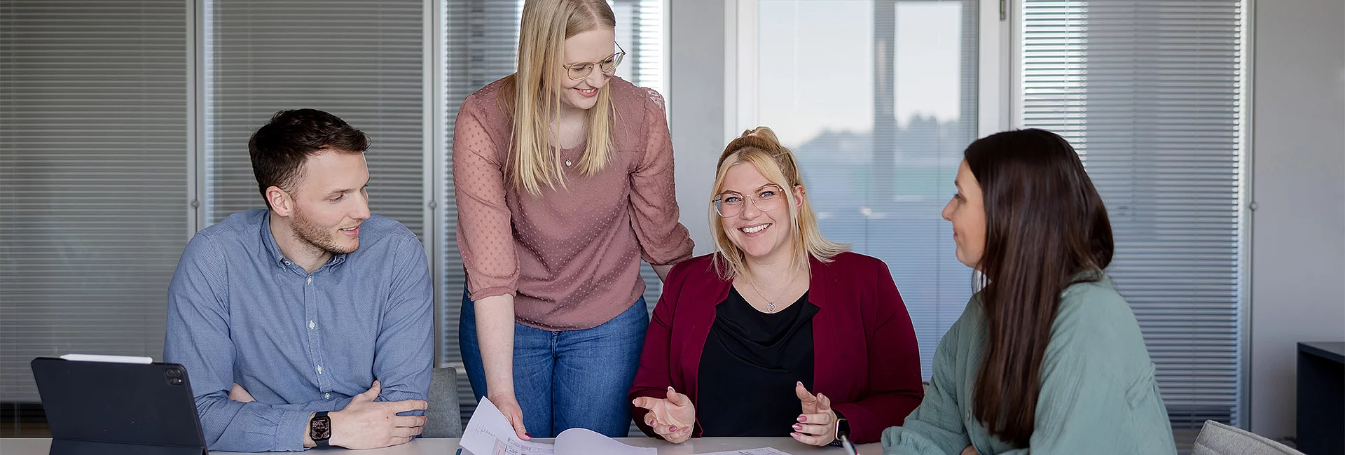 Vier Personen unterhalten sich an einem Tisch mit Papieren, während eine lächelnde Person in der Mitte sitzt. Umgebung: Büro mit Fenstern und Jalousien im Hintergrund.