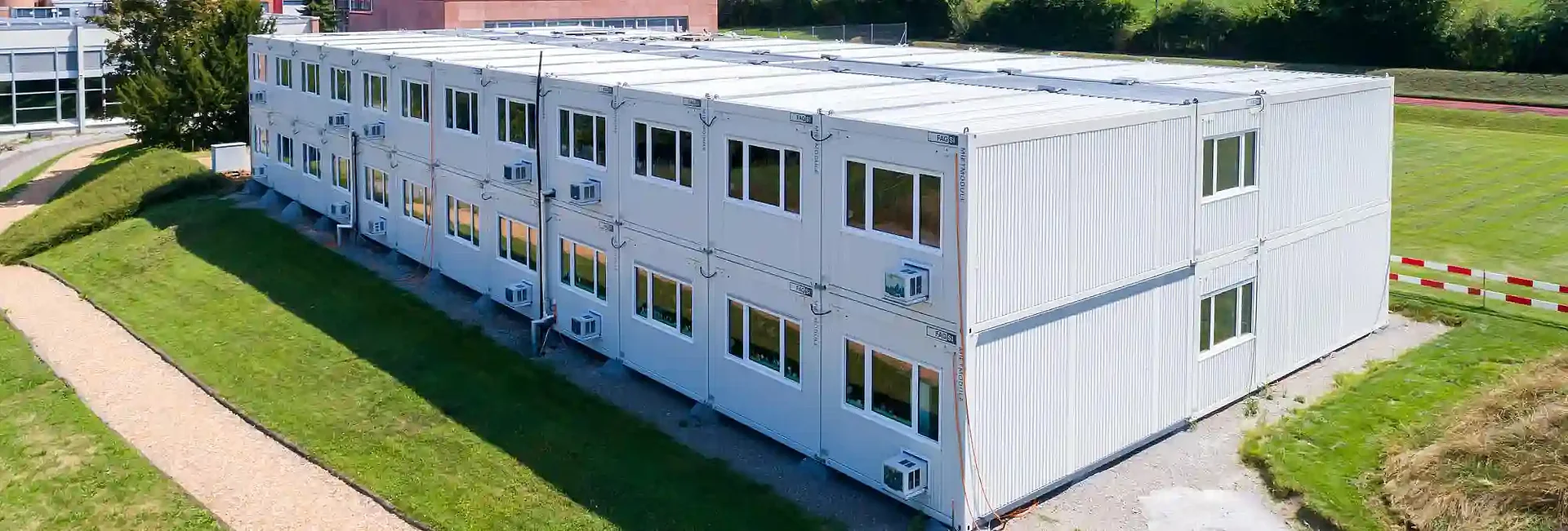 Bâtiment modulaire de deux étages composé de modules préfabriqués, implanté dans un environnement verdoyant. Le bâtiment clair dispose de nombreuses fenêtres et d’équipements techniques extérieurs, adaptés à des usages de bureaux, d’administration ou d’hébergement.