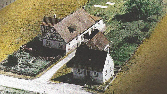 Zwei nebeneinander stehende Landhäuser - ein Fachwerkhaus und ein schlichtes Wohnhaus — mit Einfahrt und kleinem Garten, umgeben von Feldern und Bäumen in ländlicher, landwirtschaftlicher Umgebung.