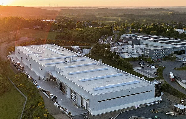 Ein großes weißes Industriegebäude mit flachem Dach und Dachfenstern steht im Vordergrund; es ist von Parkplätzen, Zufahrtswegen und weiteren Fabrikgebäuden umgeben, im Hintergrund Hügel bei Sonnenuntergang.