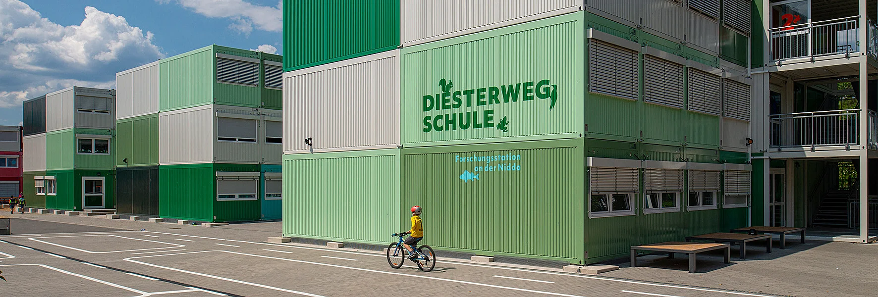 Kind mit Fahrrad vor grünen modularen Schulcontainern der „Diesterwegschule“ auf einem sonnigen Schulhof.