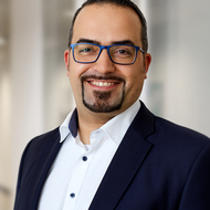 Ein lächelnder Mann mit blauer Brille und kurzem Bart trägt weißes Hemd und dunkles Sakko, steht frontal und schaut in die Kamera; unscharfer, heller Innenraum im Hintergrund.