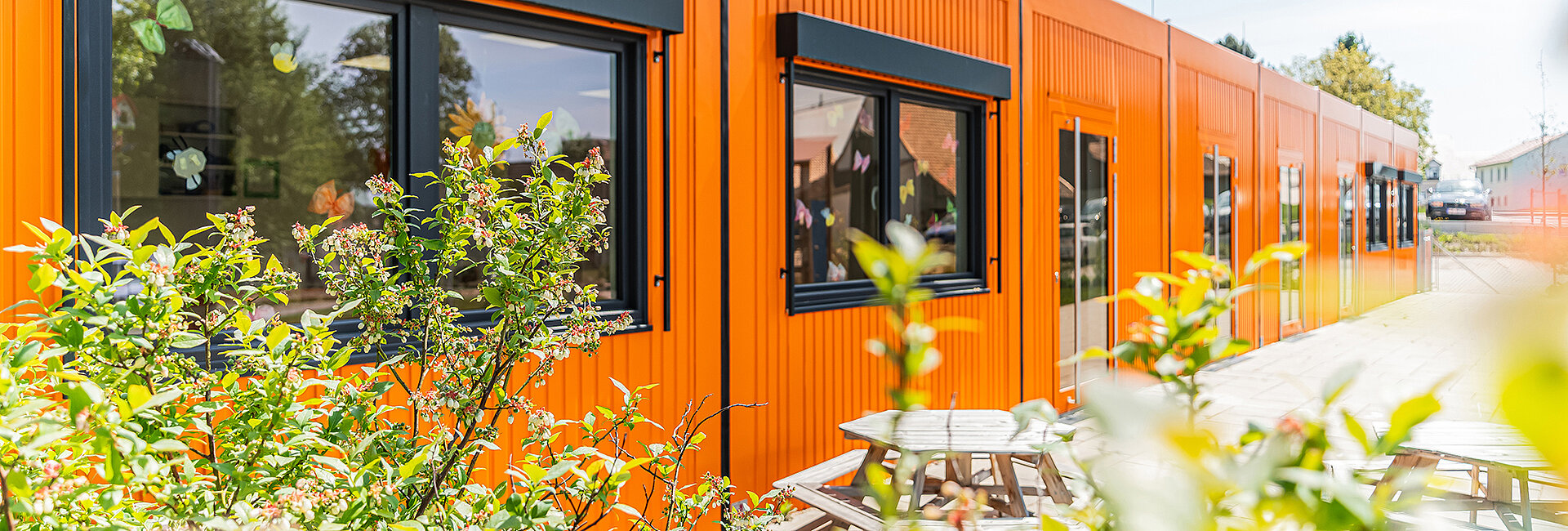 Orange Modulgebäude steht längs; Fenster zeigen bunte Dekorationen. Im Vordergrund blühen Sträucher, davor stehen Holzbänke/Picknicktische auf gepflastertem Weg; helle, sonnige Außenanlage.