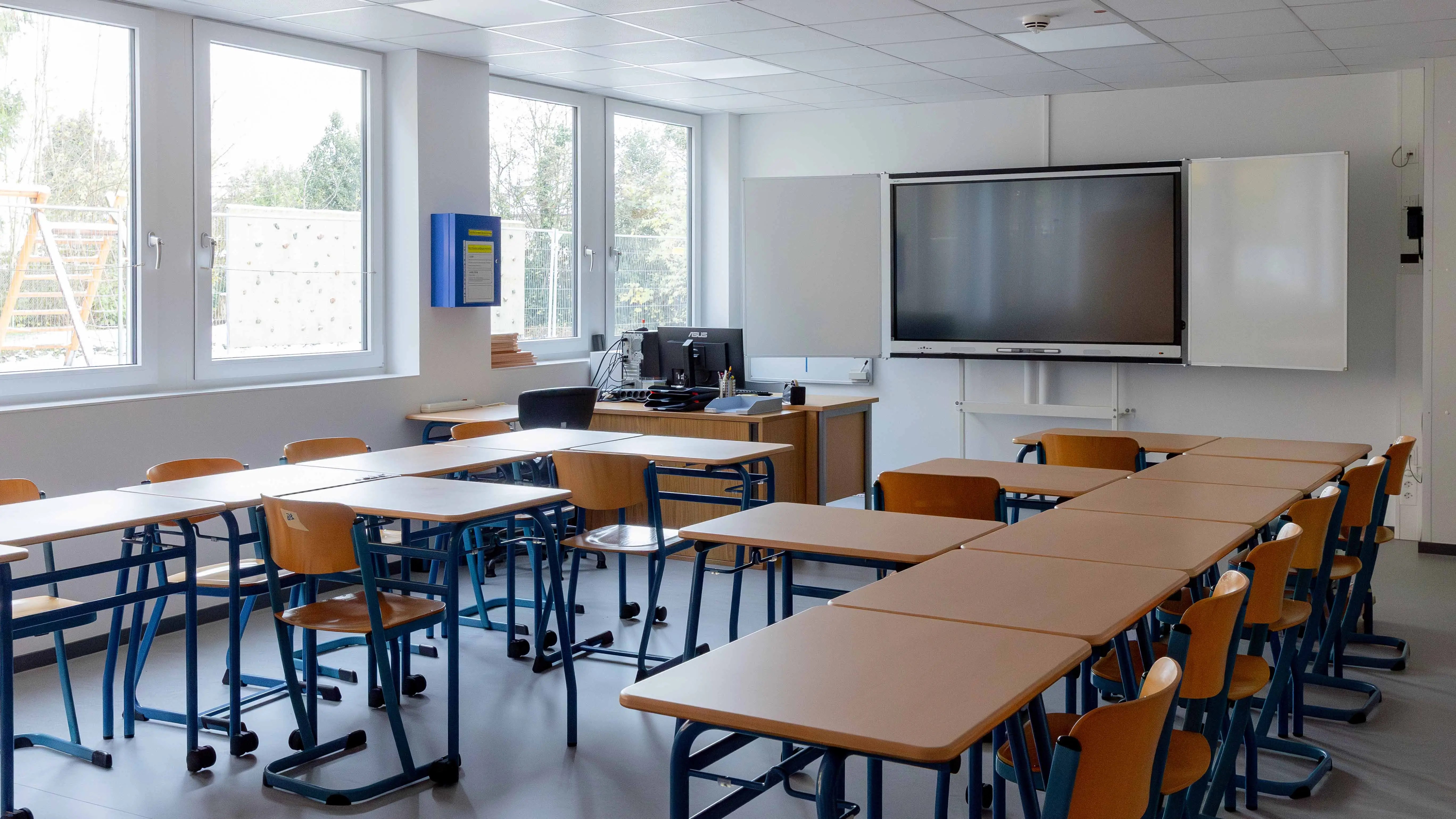 Unterrichtsraum mit Tischen, Stühlen, großer digitaler Tafel und Fenstern zum Spielplatz
