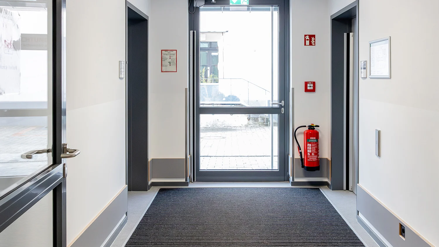 Heller Eingangsflur mit Glastür, Aufzug, Feuerlöscher und dunkler Schmutzfangmatte