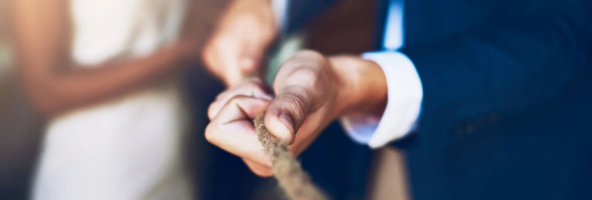 Eine Hand in einem Anzug hält ein Seil fest, zieht es energisch, während eine verschwommene Person im Hintergrund zu sehen ist.