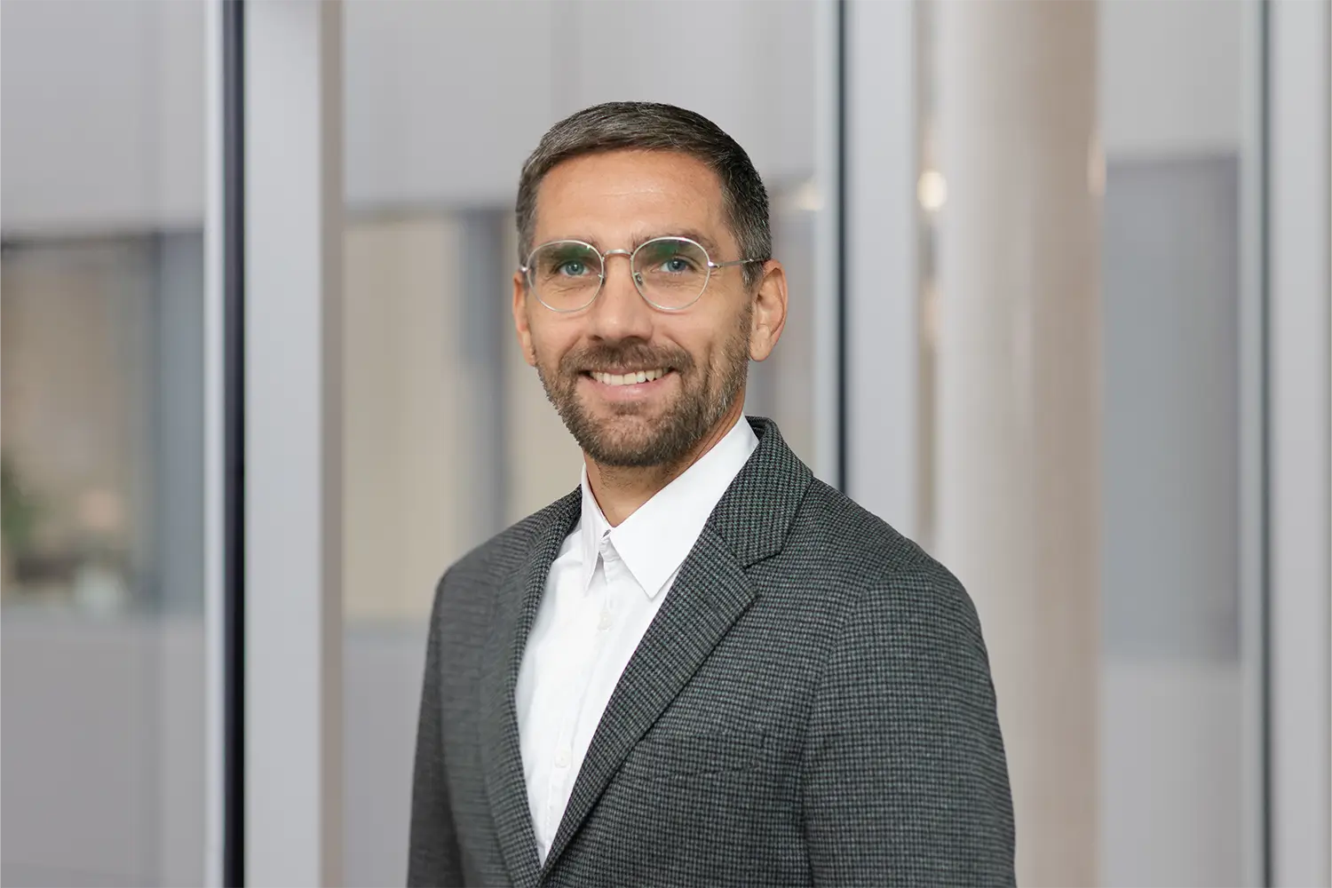 Porträt von Benjamin Hien mit runder Brille und grauem Tweed-Sakko vor einer Glasfront