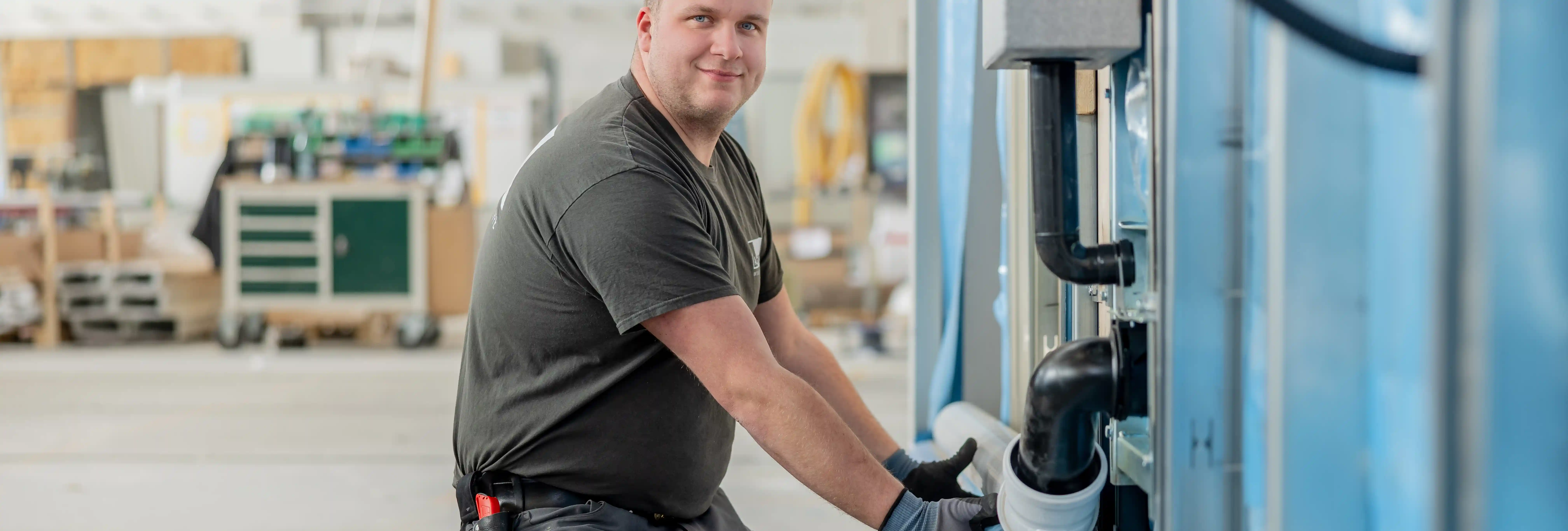 Eine Person mit Arbeitshandschuhen arbeitet in einer hellen Produktionshalle an einer großen blauen Maschine. Die Person befestigt oder hält ein weißes Rohr, das mit schwarzen Rohrleitungen verbunden ist. Im unscharfen Hintergrund sind Werkstatt- und Industrieeinrichtungen zu erkennen.