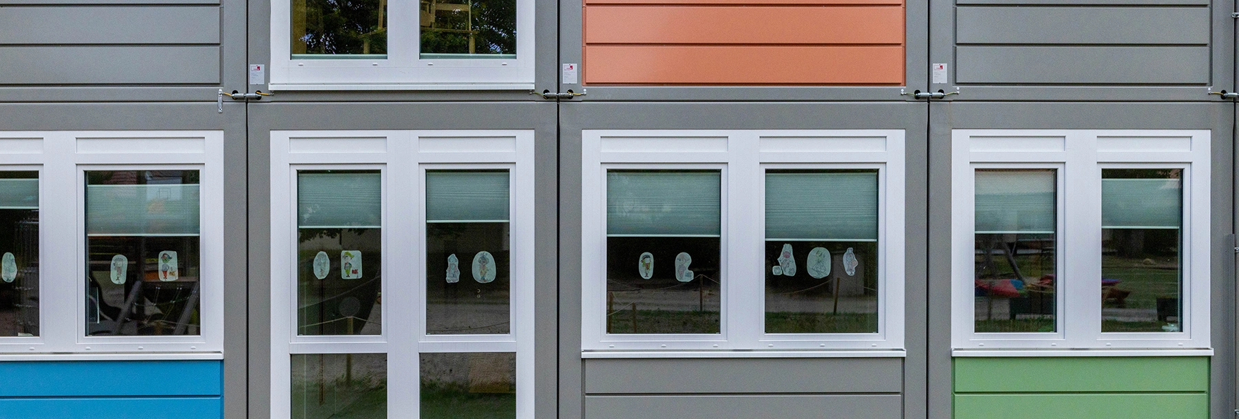 Mehrere weiße Fenster eines bunten Modulgebäudes zeigen an den Scheiben angeklebte Kinderzeichnungen; halb heruntergelassene Jalousien, reflektierte Außenflächen und ein Spielplatz im Hintergrund.
