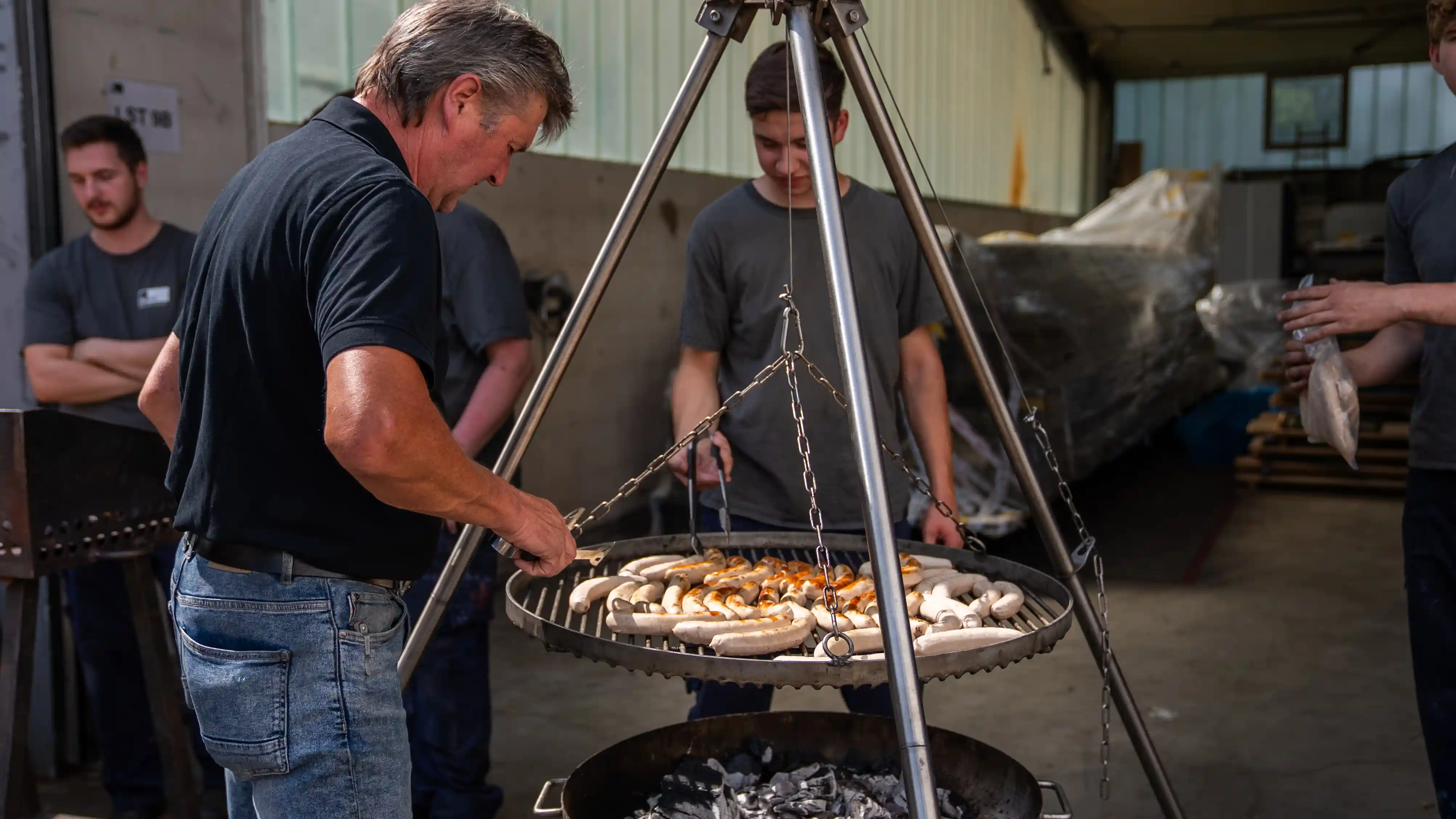 Ein Ausbilder grillt Bratwürste auf einem Schwenk-Grill, während Auszubildende in der Werkshalle zusehen