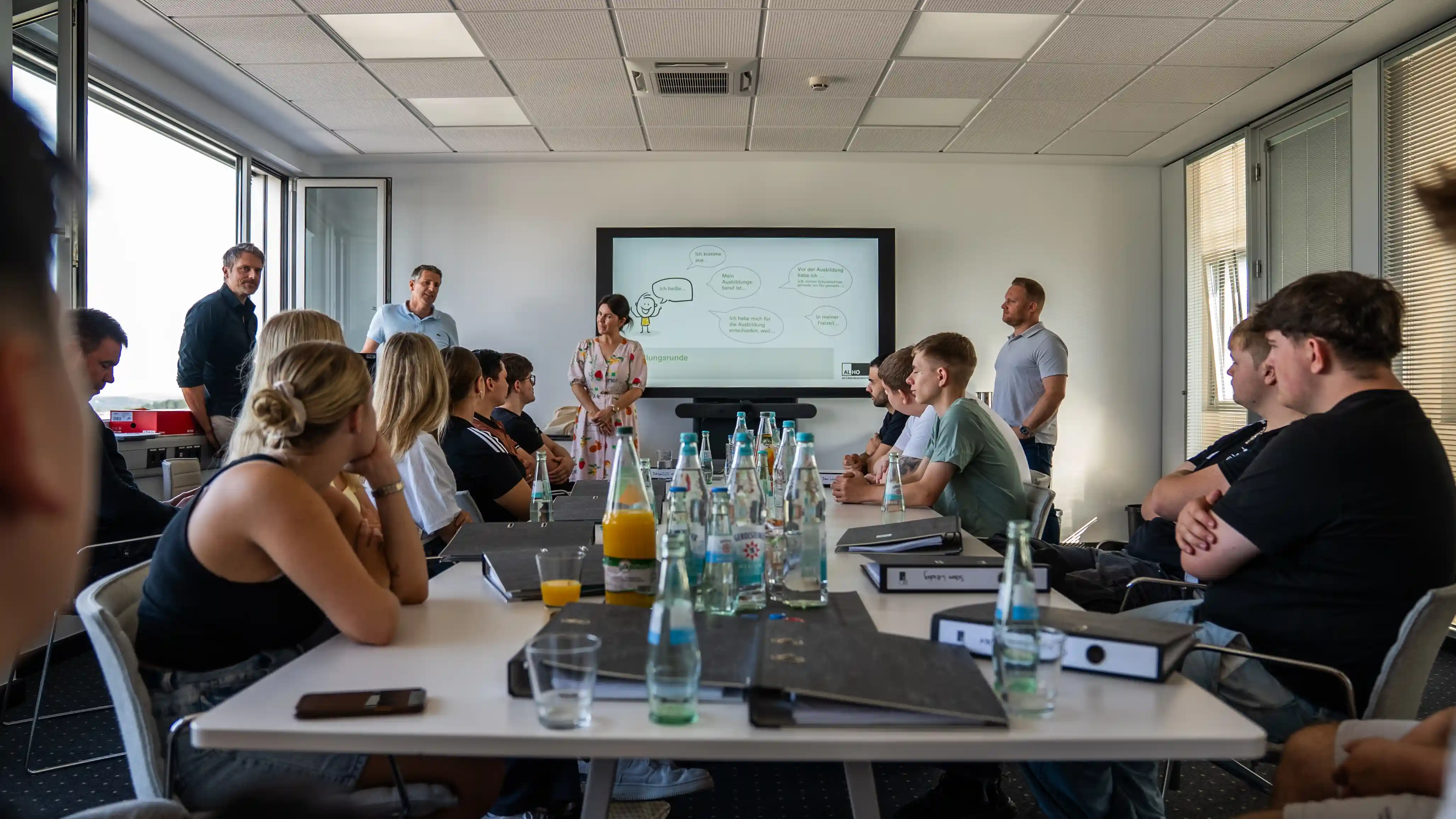 ALHO Mitarbeitende und Auszubildende sitzen gemeinsam in einem Besprechungsraum bei einer Vorstellungsrunde mit Getränken auf dem Tisch