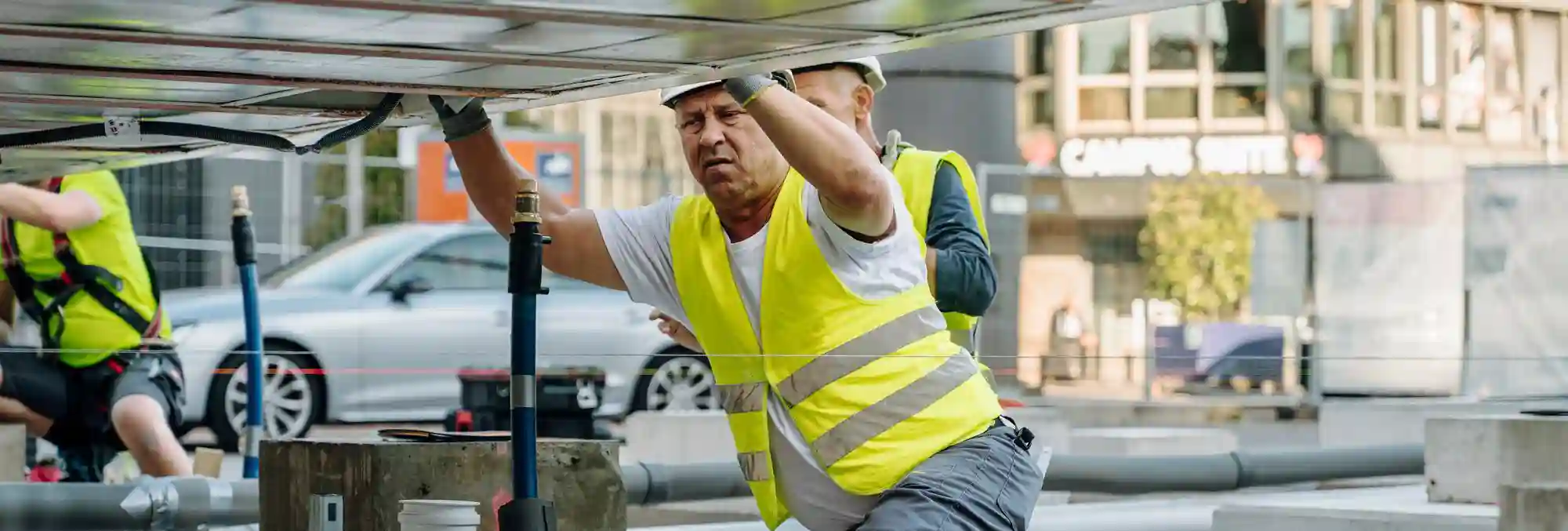 Personne portant un gilet de haute visibilité jaune et un casque de sécurité, travaillant sur une structure métallique sur un chantier. D’autres éléments de construction et zones de travail sont visibles à l’arrière-plan. L’image illustre un environnement professionnel lié aux travaux de construction et de montage.
