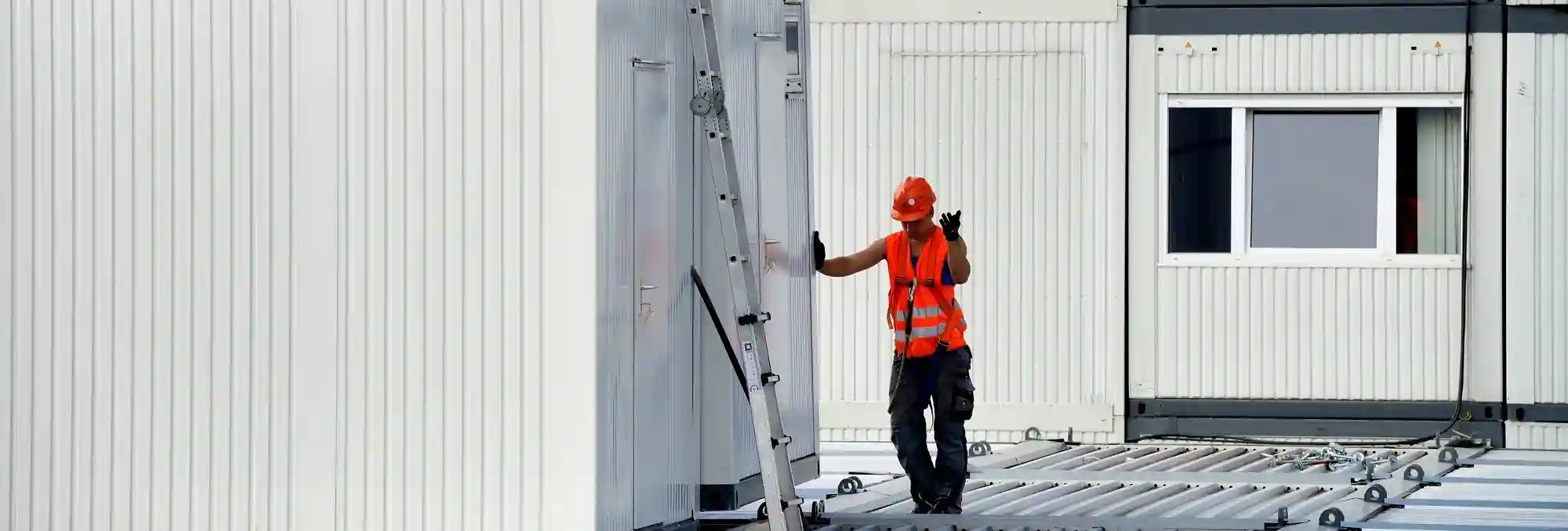 echnicien intervenant sur l’extension d’un bâtiment modulaire, équipé d’un gilet haute visibilité et d’un casque de sécurité. Des modules préfabriqués sont visibles en arrière-plan, illustrant les travaux d’assemblage et de montage sur site.