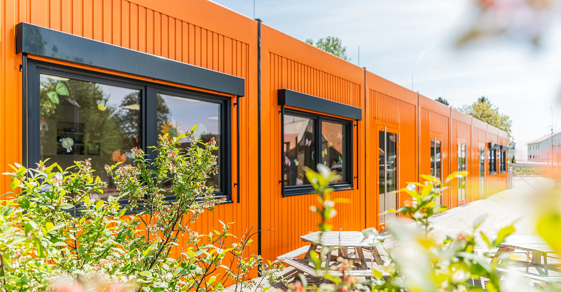 Außenansicht einer modernen Kindertagesstätte von FAGSI in modularer Bauweise. Das eingeschossige Kita-Gebäude mit orangefarbener Fassade, großen Fenstern und begrüntem Außenbereich bietet eine freundliche und funktionale Lern- und Spielumgebung für Kinder.