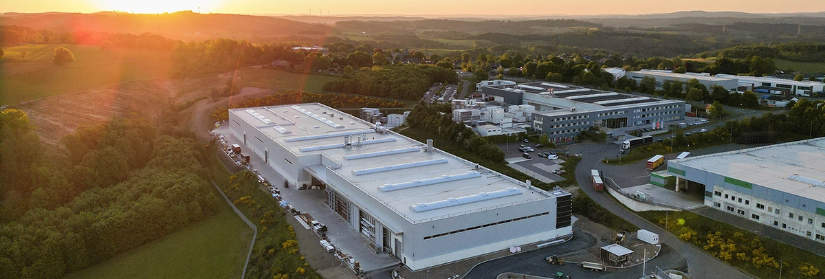 Ein großes weißes Industriegebäude mit flachem Dach und Dachfenstern steht im Vordergrund; es ist von Parkplätzen, Zufahrtswegen und weiteren Fabrikgebäuden umgeben, im Hintergrund Hügel bei Sonnenuntergang.