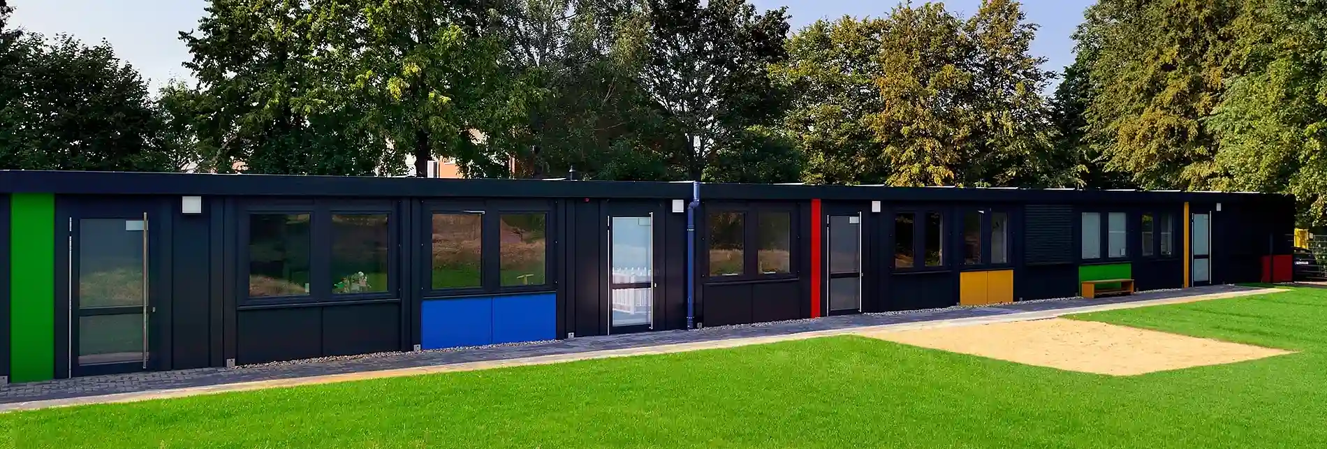 Barrierefreier, eingeschossiger Modulbau mit dunkler Fassadengestaltung und farbigen Akzenten. Vor dem Gebäude befinden sich eine gepflegte Rasenfläche und ein Gehweg, während hohe Bäume die Anlage umrahmen.