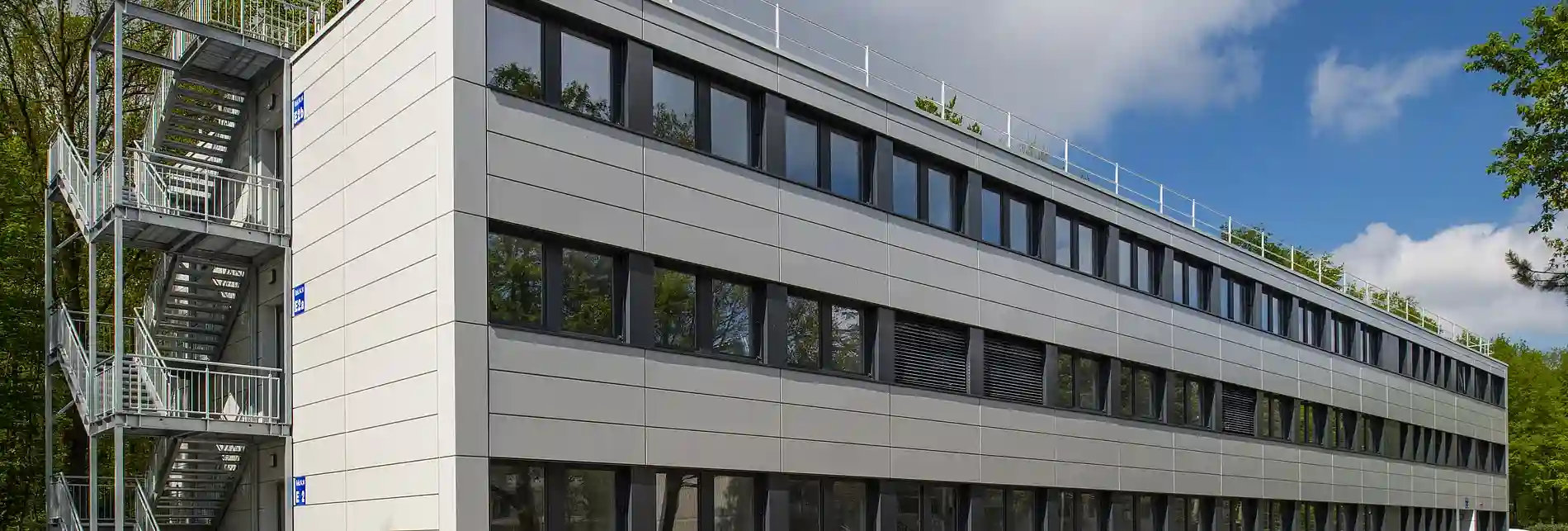Dreigeschossiges modulares Büro- oder Verwaltungsgebäude mit heller Fassadenverkleidung, großen Fensterbändern und außenliegendem Treppenturm. Das Gebäude ist von Grünflächen und Bäumen umgeben und zeigt eine moderne, funktionale Architektur.