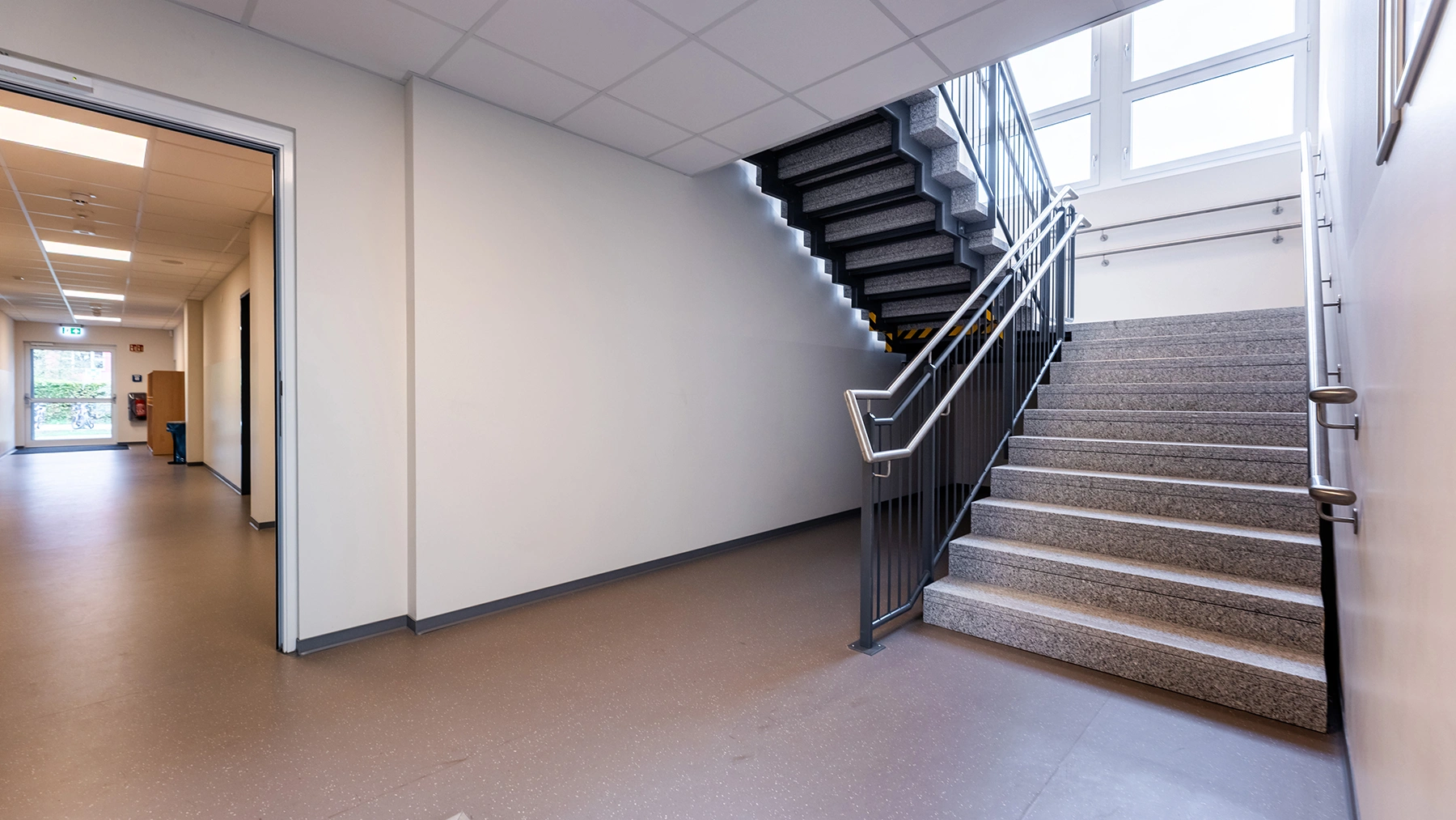 Helles Treppenhaus mit grauer Steintreppe, Metallgeländer und angrenzendem Flur