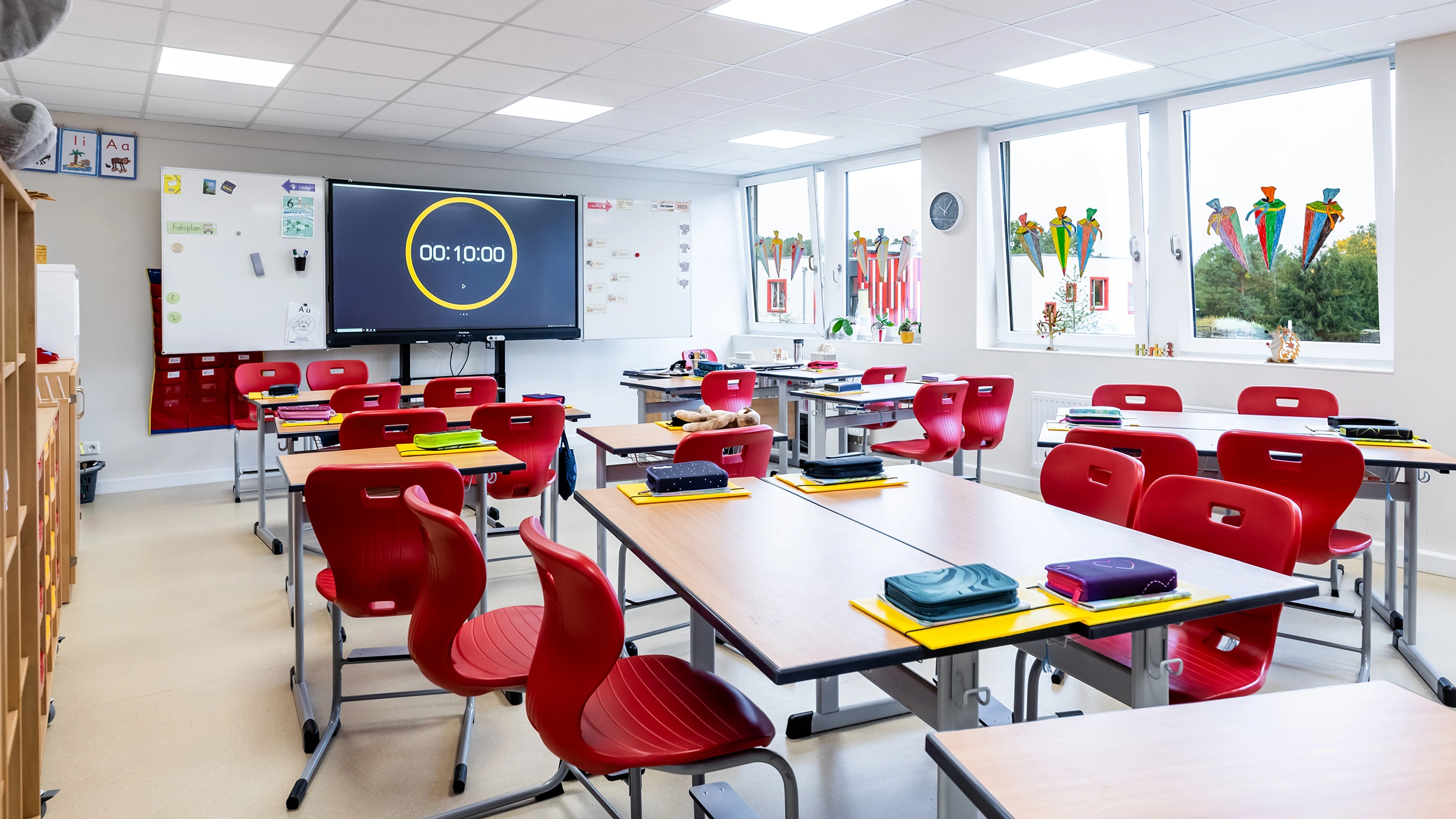 Klassenzimmer mit roten Stühlen, Schultischen, digitaler Tafel und bunten Fensterdekorationen