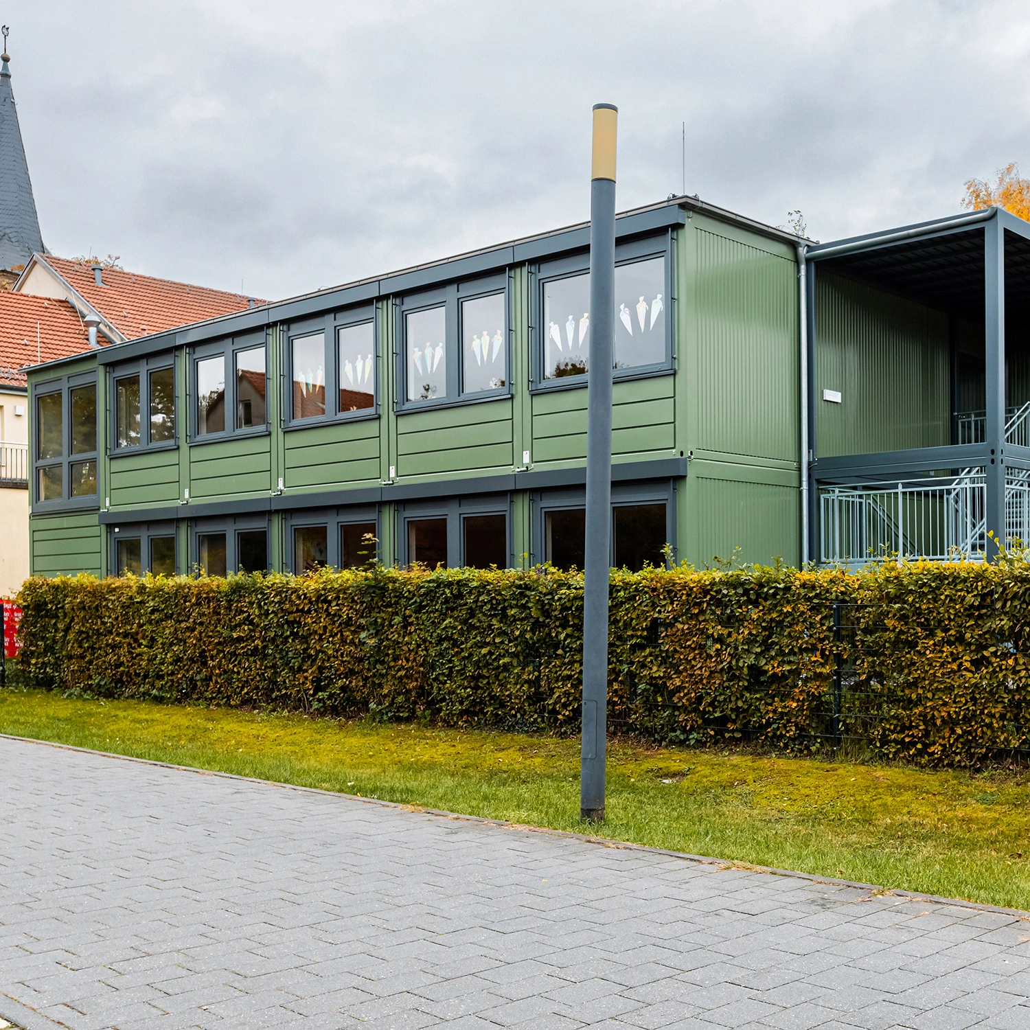 Grünes zweigeschossiges Modulgebäude mit Fensterreihen, Außentreppe und Hecke an der Straße