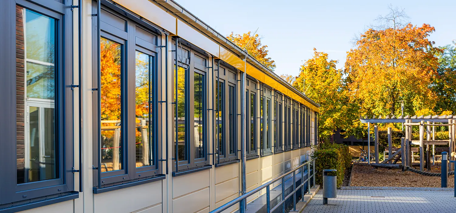 Lange Fensterreihe einer niedrigen Gebäudefassade, die gold-orange Herbstbäume spiegelt; das Gebäude steht am gepflasterten Weg neben einem hölzernen Spielplatz unter klarem blauem Himmel.