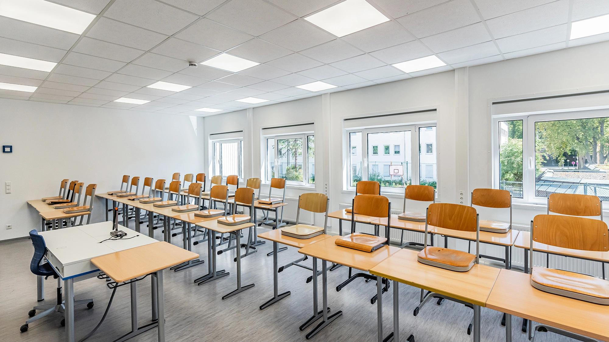 Klassenzimmer mit aufgestellten Stühlen, Lehrerpult, großen Fenstern und heller Deckenbeleuchtung