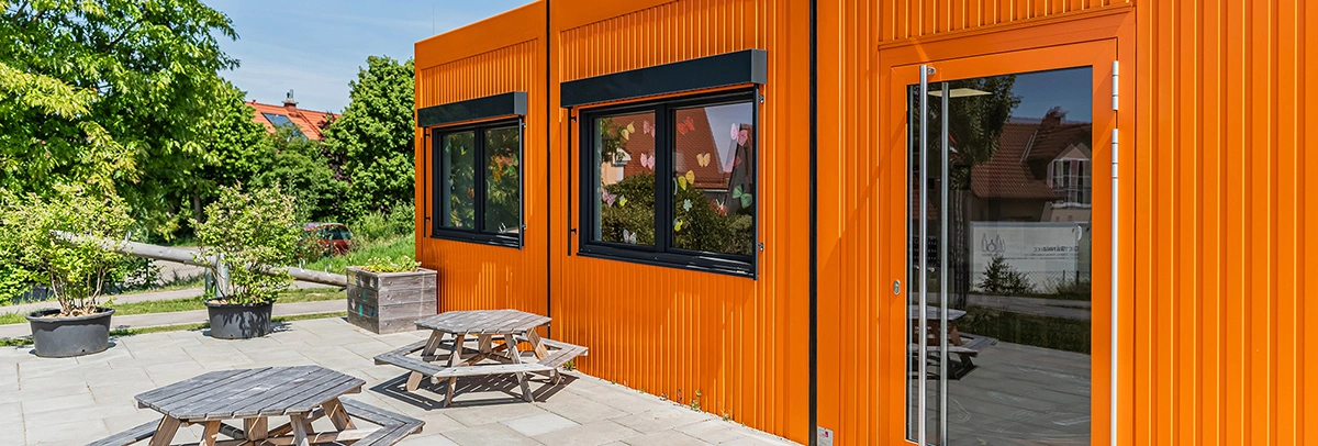 Ein orangefarbener Modulbau steht in einem sonnigen Innenhof; Fenster mit Papier-Schmetterlingen, davor Holz-Picknicktische und Pflanzkübel, im Hintergrund Bäume und Wohnhäuser.