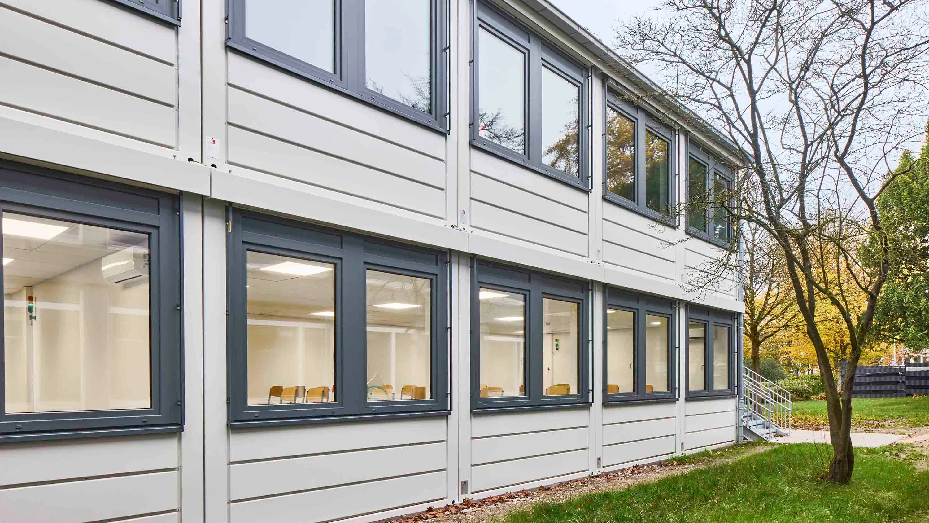 Zweigeschossige helle Containeranlage mit Fensterreihen, Außentreppe und Grünfläche