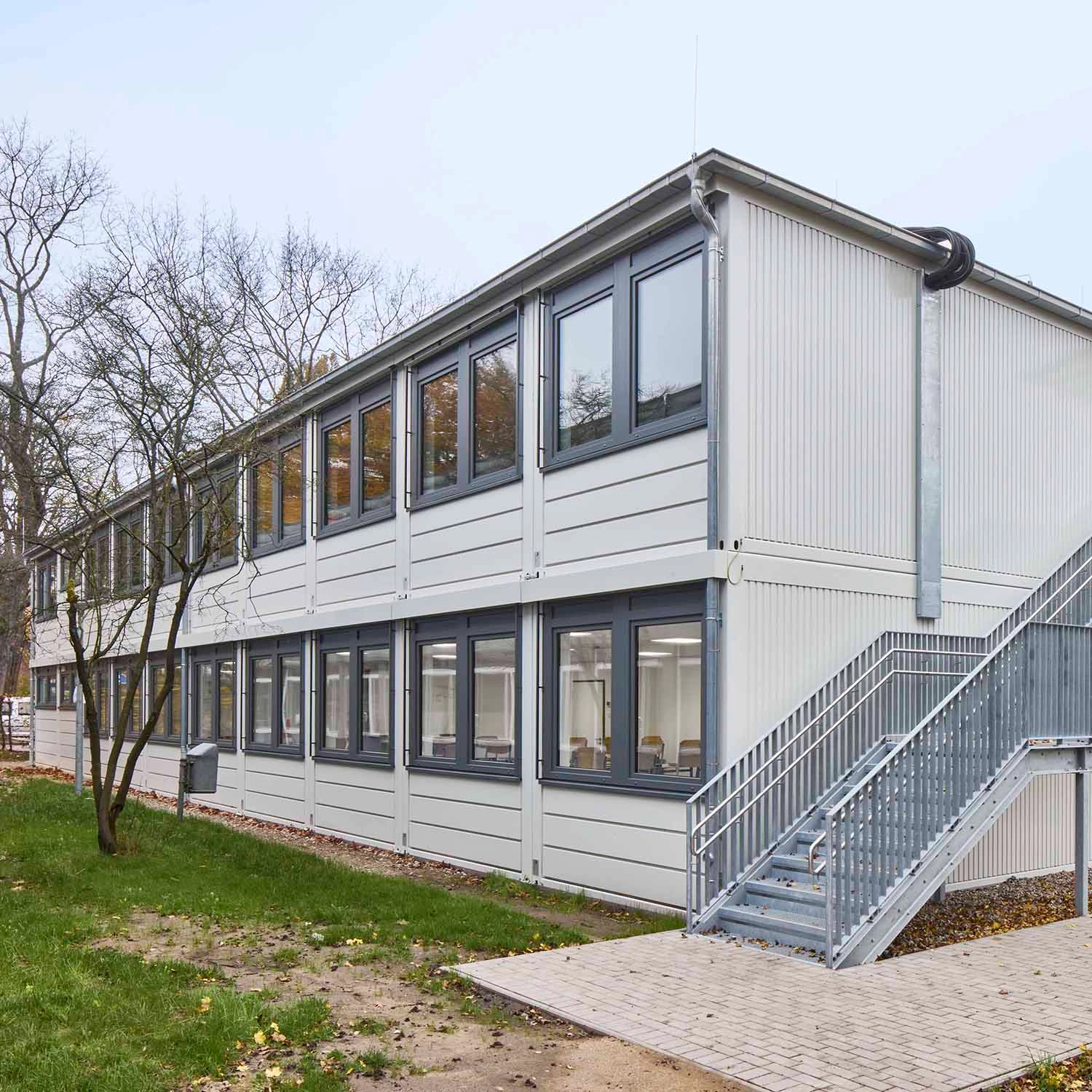 Zweigeschossige helle Containeranlage mit Außentreppe, Fensterreihen und Bäumen im Außenbereich
