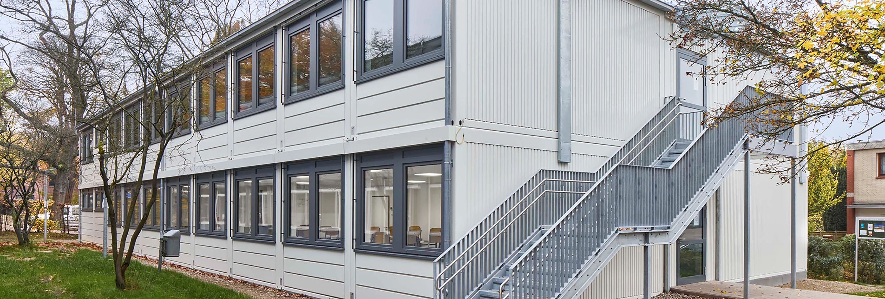 Zweigeschossige helle Containeranlage mit Außentreppe, Fensterreihen und Bäumen im Außenbereich