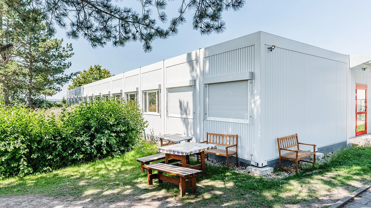 Außenbereich eines Hortgebäudes mit Sitzbänken, Holztisch und weißer Fassade