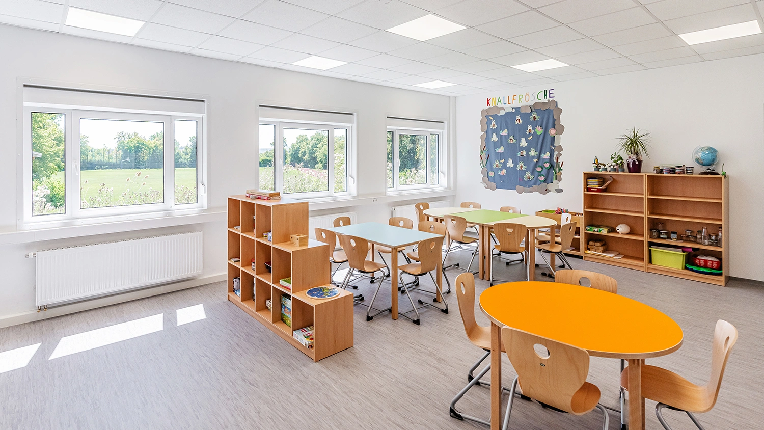 Heller Hortgruppenraum mit Kindertischen, Holzstühlen, Regalen und großer Fensterfront