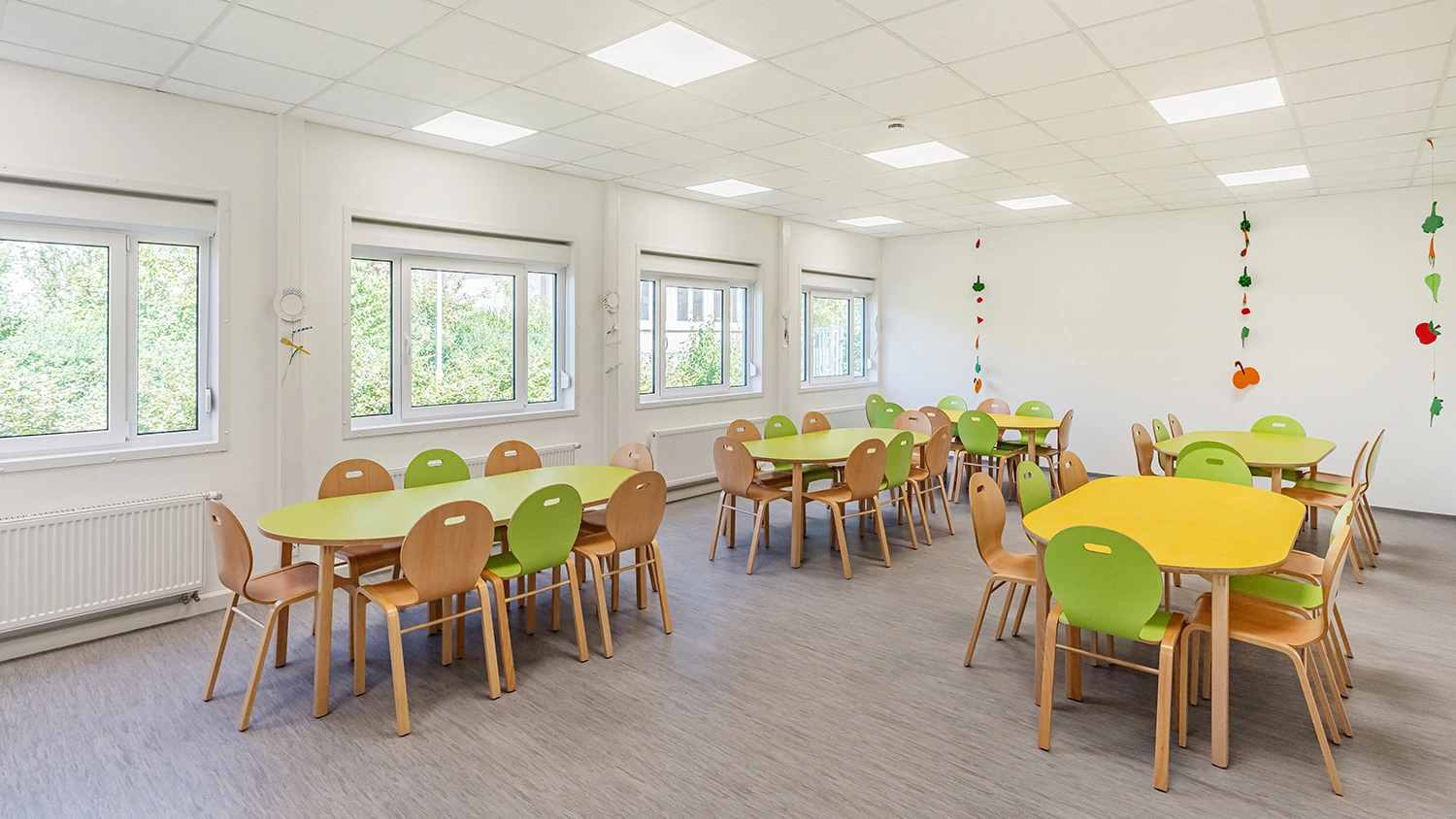 Heller Gruppenraum mit Kindertischen, grünen Stühlen und großen Fenstern