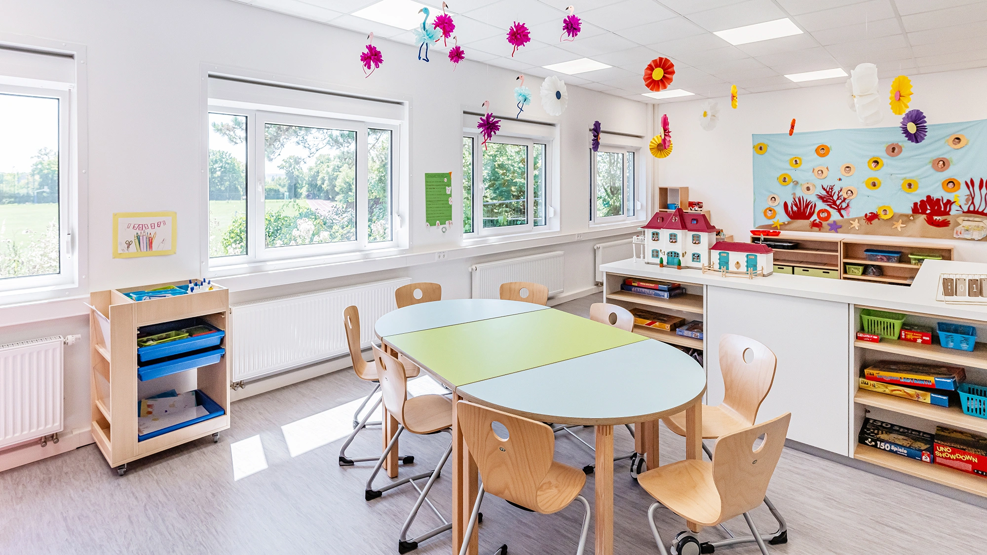 Heller Gruppenraum mit Kindertischen, Holzstühlen, Spielregalen und bunter Wanddekoration