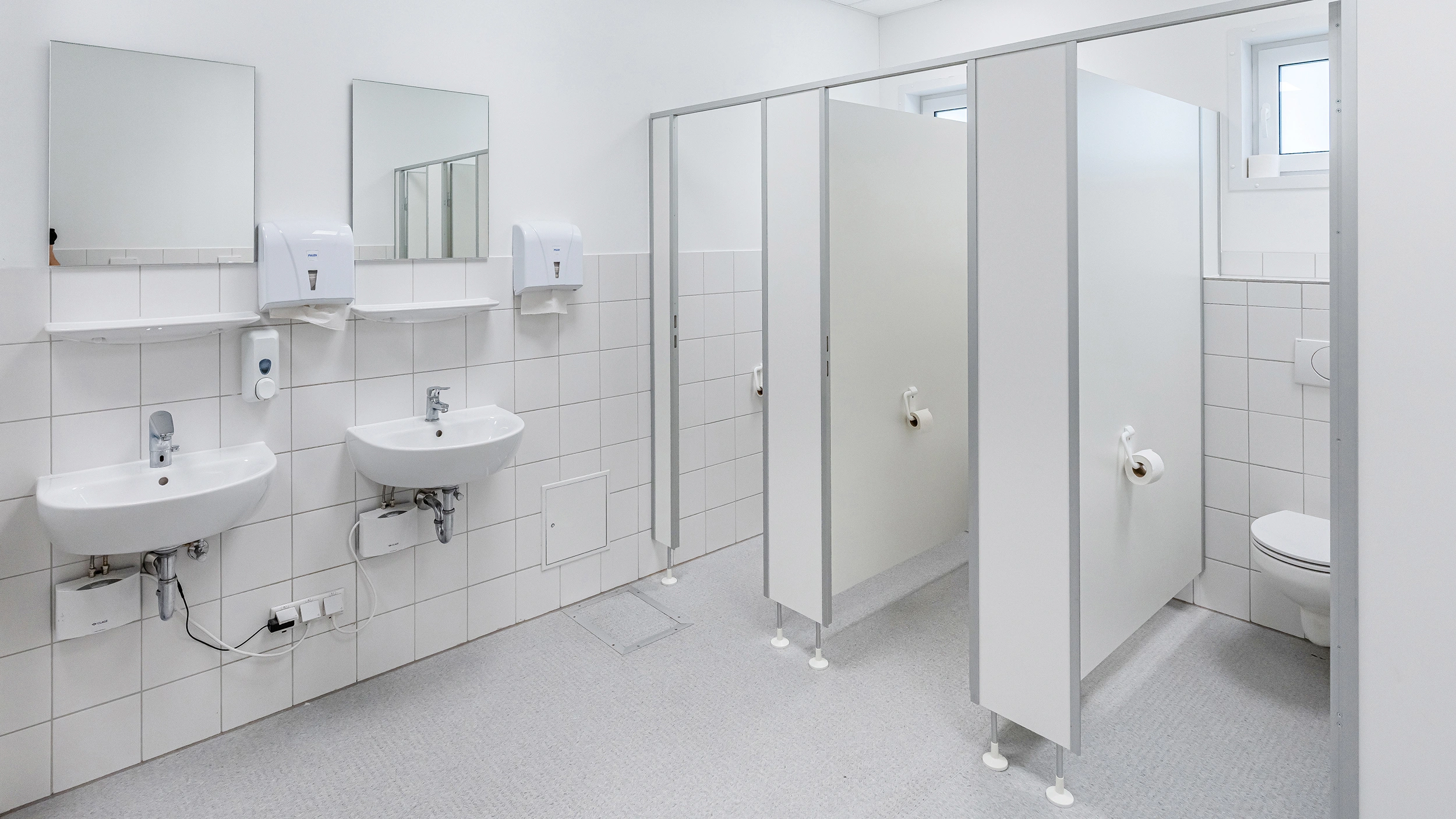 Sanitärraum mit zwei Waschbecken, Spiegeln, Papierhandtuchspendern und WC-Kabinen