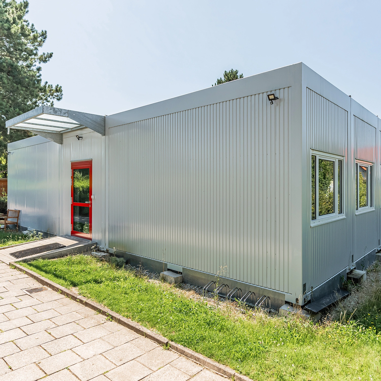 Eingangsbereich eines hellen Hortgebäudes in Containerbauweise mit roter Tür und Glasvordach