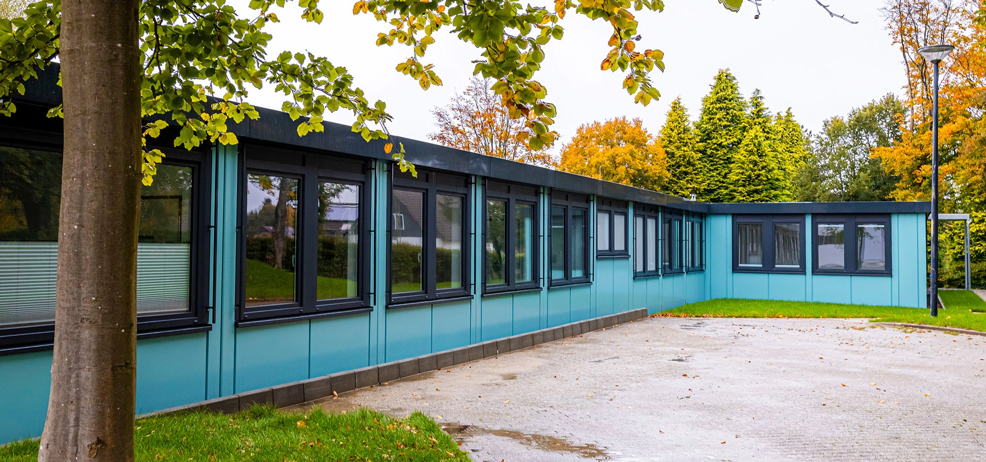 Einstöckiges Modulgebäude mit türkisfarbener Fassade und schwarzen Fensterrahmen, umgeben von Bäumen mit herbstlichem Laub, davor gepflasterte Fläche und Rasen.
