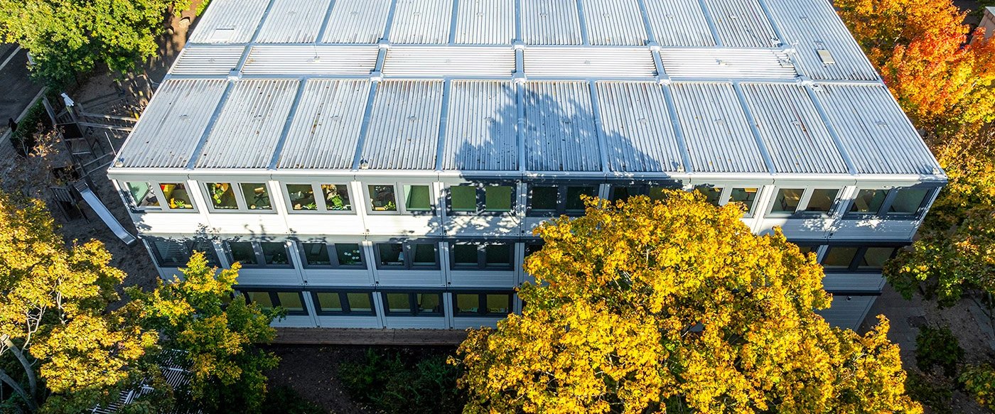 Mehrgeschossiger modularer Schul- oder Bürocontainer steht zentral; Fenster mit Pflanzen, Baumschatten fällt auf das wellige Blechdach; umgeben von herbstlich gelben Bäumen und einem kleinen Spielplatz mit Rutsche.