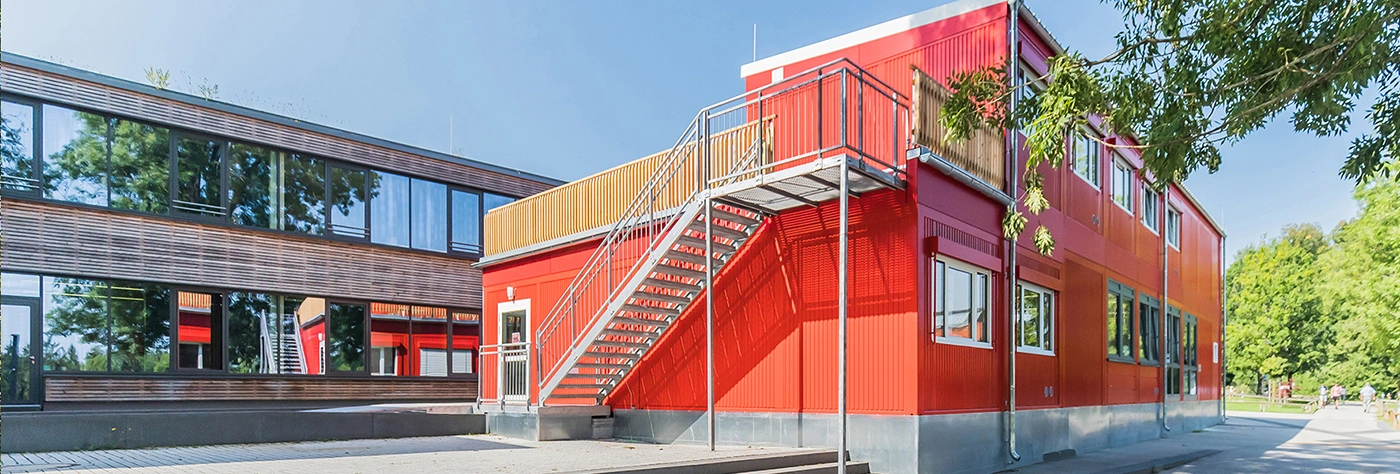 Rotes zweistöckiges Modulgebäude mit außen angebrachter Metalltreppe; Treppe führt zur oberen Ebene; im Hintergrund links langes Holzfassaden-Gebäude mit Spiegelglas, rechts Gehweg und grüner Park mit Bäumen.