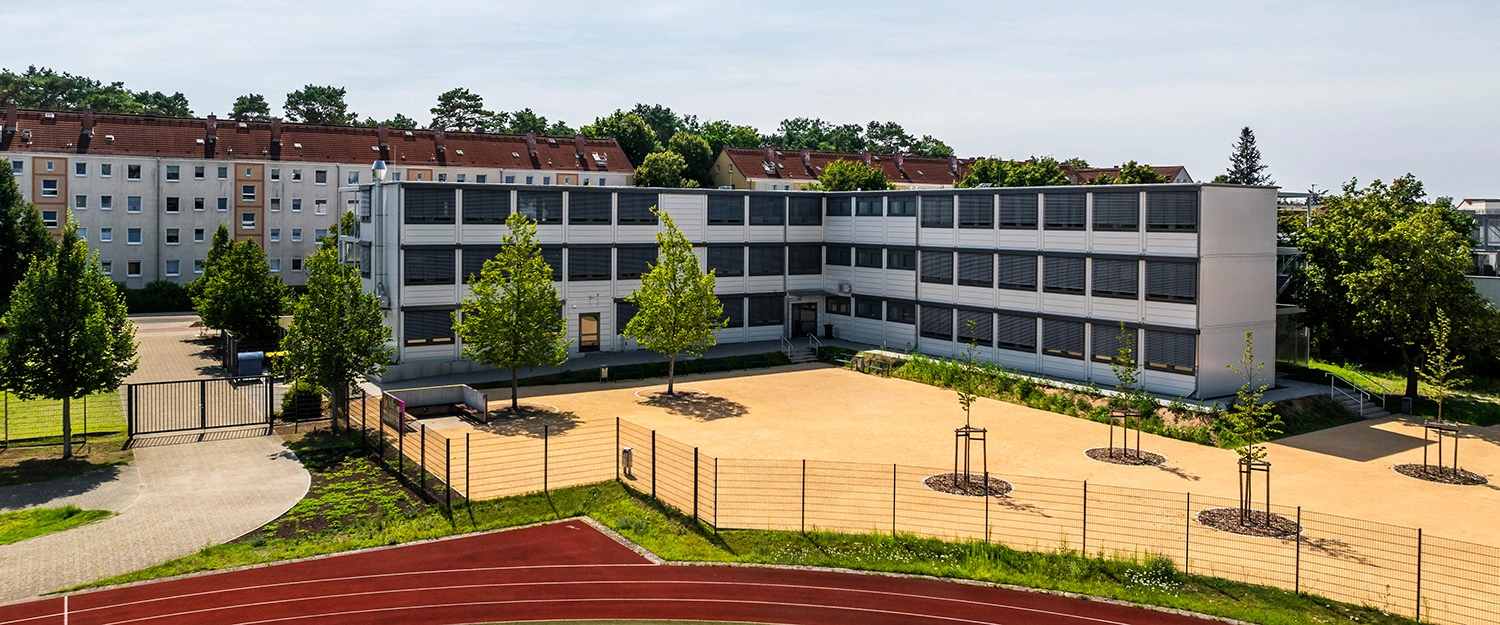 Mehrstöckiges Schulgebäude in modularer Bauweise mit Innenhof, Bäumen und eingezäunter Fläche, aufgenommen aus erhöhter Perspektive.
