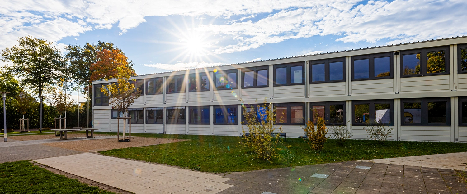 Zweigeschossiges Modulgebäude mit heller Fassade und großen Fensterreihen, Außenanlage mit Wegen, Grünflächen und Bäumen, Sonne mit Lensflare über dem Gebäude.
