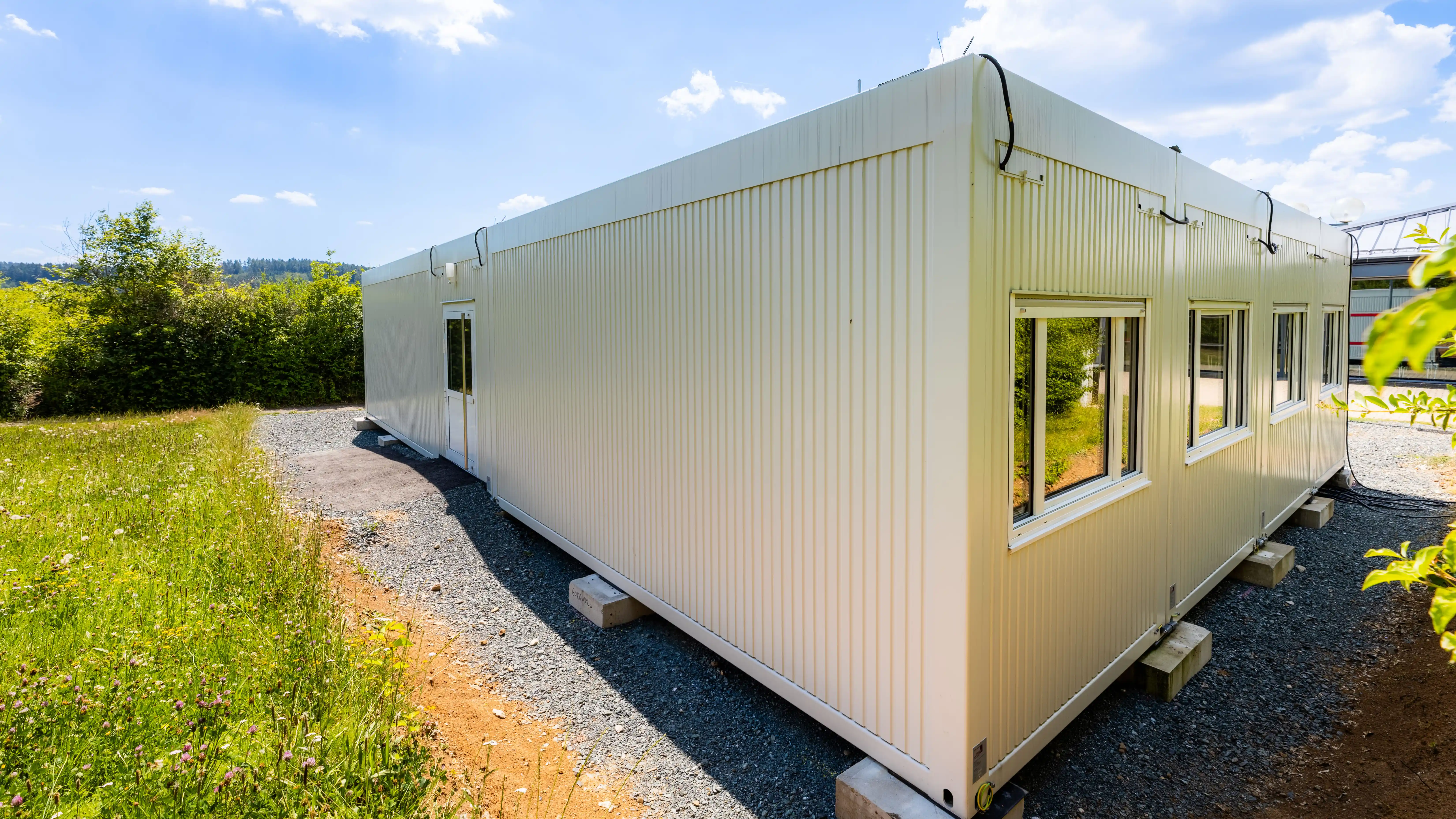 Eingeschossige ProENERGY-Containeranlage der Pestalozzi-Schule in Kronach mit heller Fassadengestaltung und großen Fensterflächen. Die Anlage steht auf einem Schotterfundament in einer grünen Umgebung.
