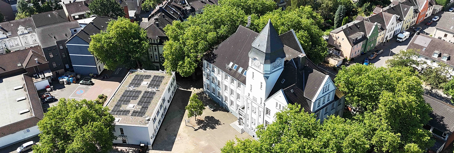 Ein großes weißes historisches Gebäude mit Turm steht auf einem Innenhof; umgeben von grünen Bäumen, daneben ein flaches Gebäude mit Solarpaneelen, im Hintergrund Wohnhäuser und Straßen.